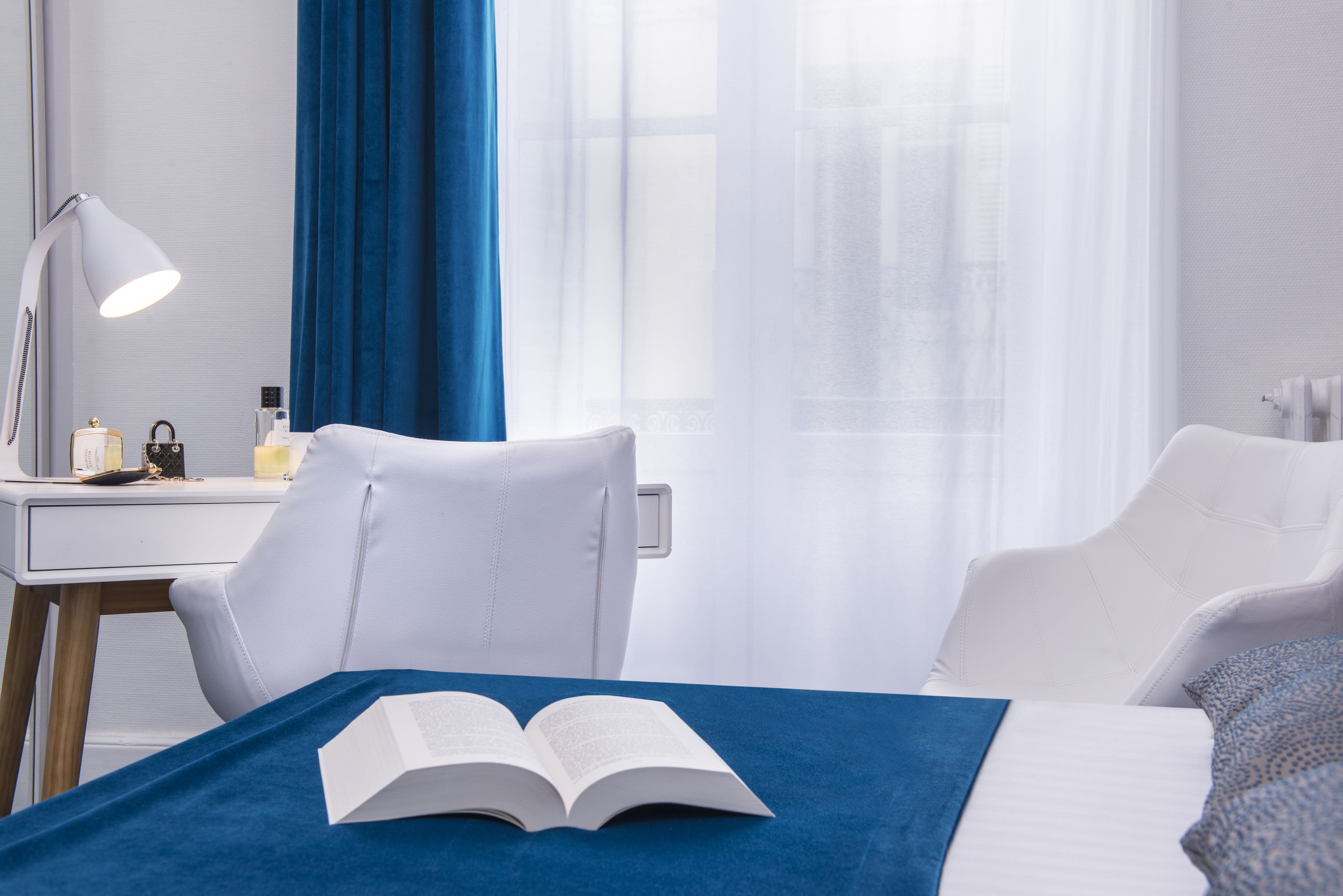 A hotel room with a bed, a desk with a lamp, a chair, and a window with sheer curtains.