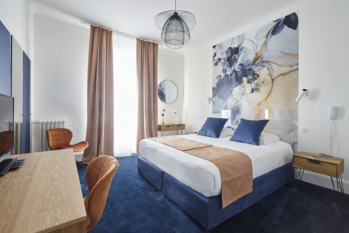 Une chambre d’hôtel avec un grand lit, un bureau avec une chaise, un miroir rond et un grand tableau abstrait accroché au mur.
