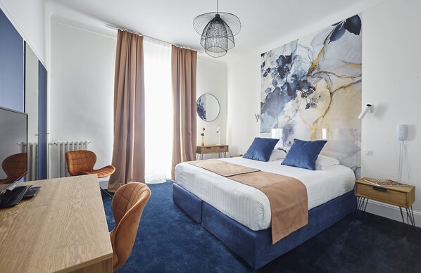 Une chambre d’hôtel avec un grand lit, un bureau avec une chaise, un miroir rond et un grand tableau abstrait accroché au mur.