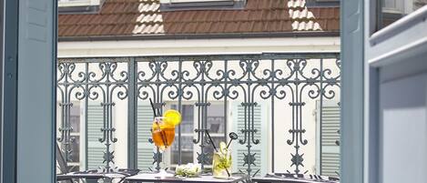 Ein Balkon mit schmiedeeisernem Geländer, einem Tisch mit Getränken und Blick auf ein Haus mit Ziegeldach und Fensterläden.