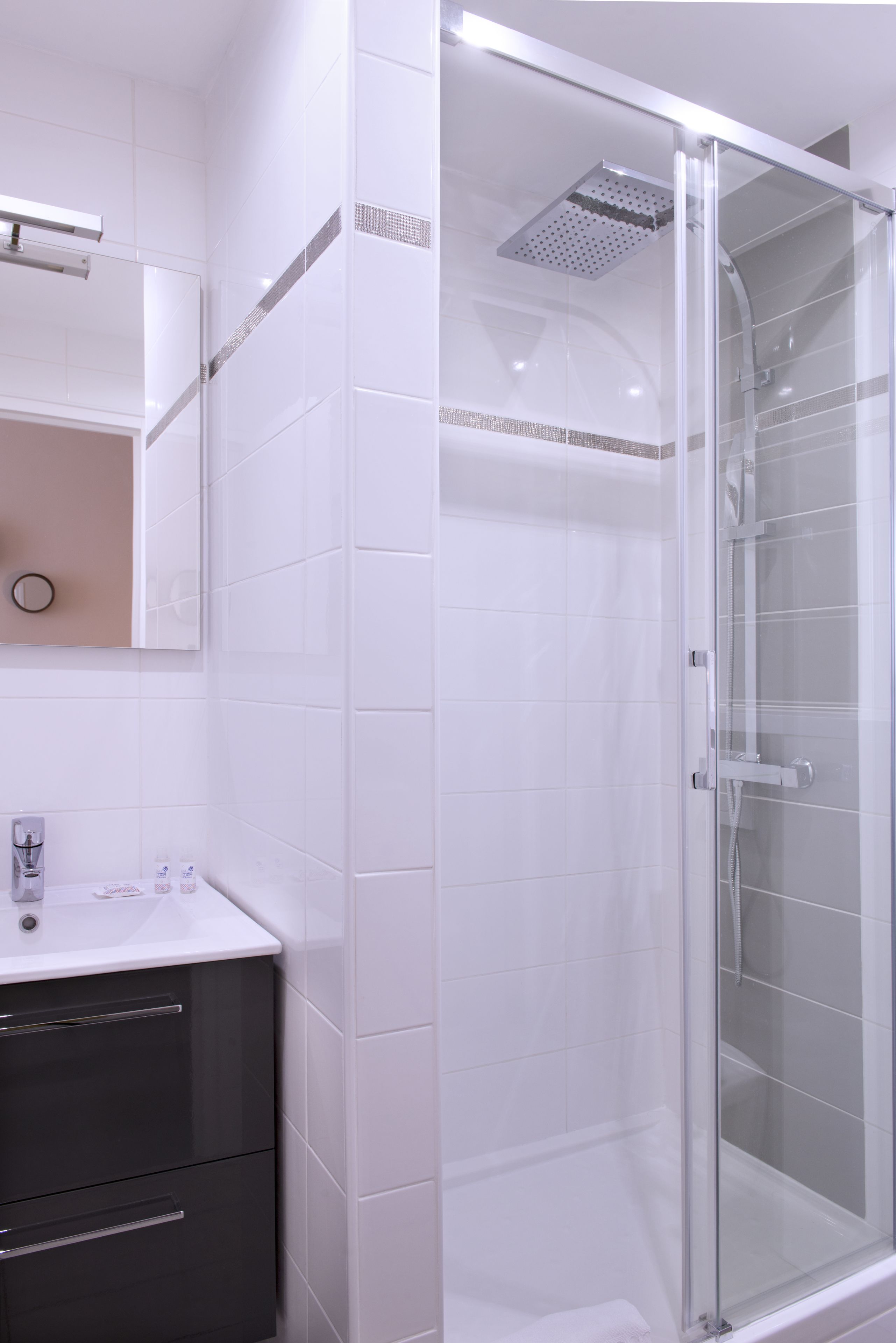 A modern bathroom with a glass-enclosed shower, white tiles, and a dark-toned vanity with drawers.