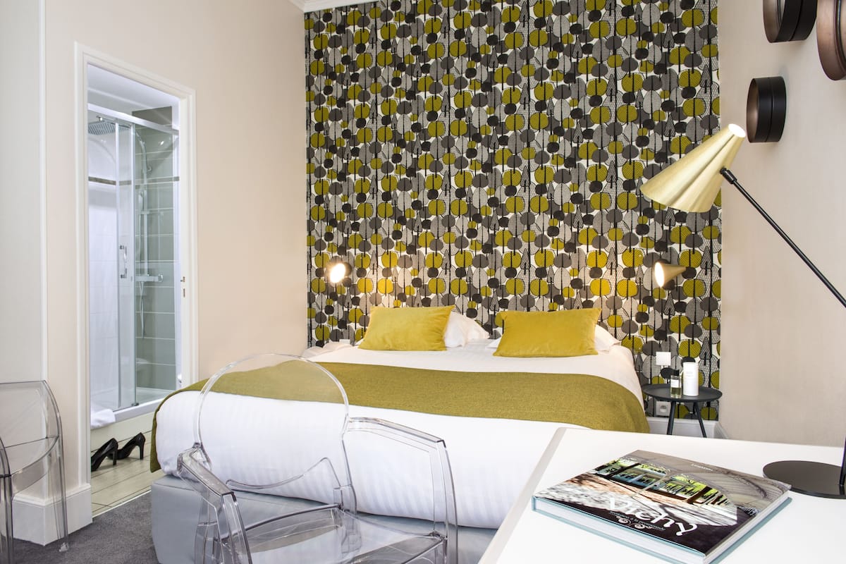 Une chambre d'hôtel moderne avec un papier peint à motifs, un lit avec une literie blanche et jaune, une table en verre transparent et un magazine posé sur la table.