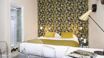 A modern hotel room with a patterned wallpaper, a bed with white and yellow bedding, a clear glass table, and a magazine on the table.