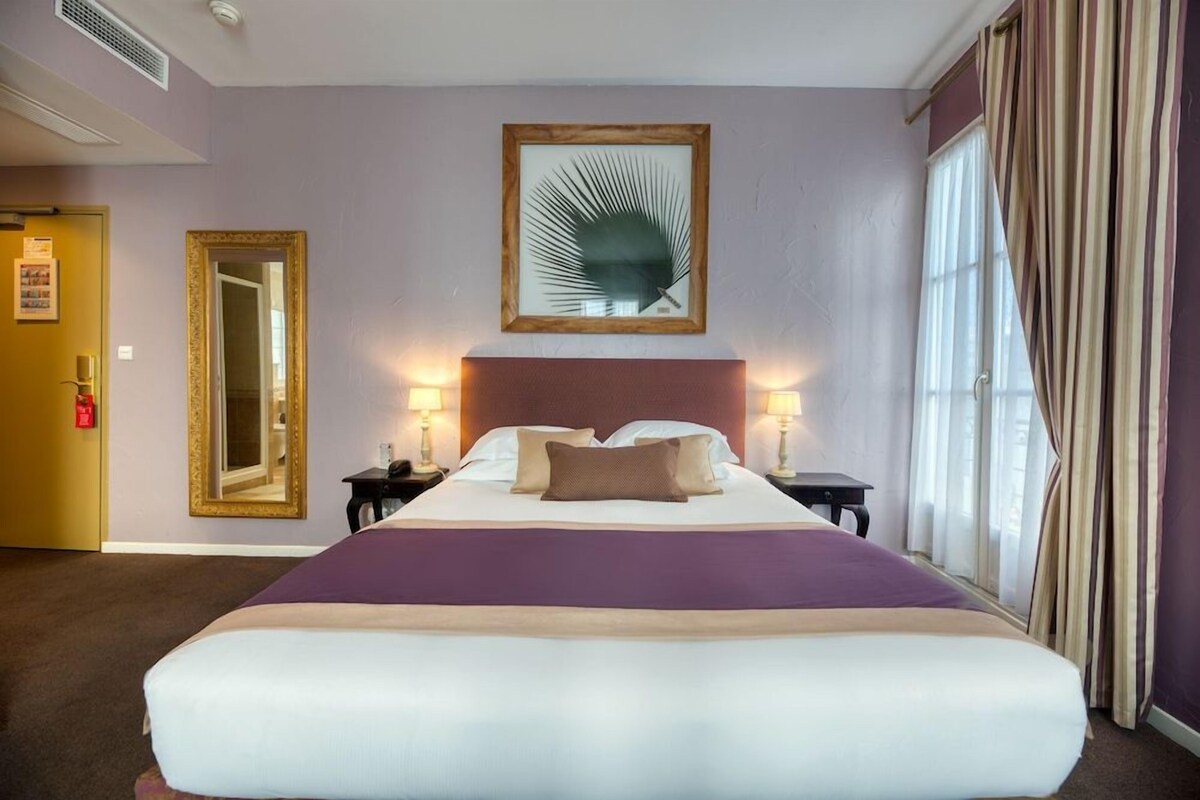 Habitación de hotel con una cama grande, un cabecero, dos mesitas de noche con lámparas, una obra de arte enmarcada en la pared y una ventana con cortinas.
