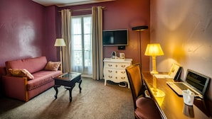 A hotel room with a red sofa, a small wooden coffee table, a desk with a laptop, a TV, and a bedside lamp.