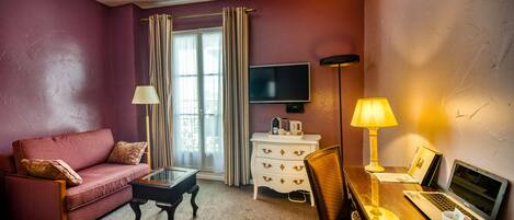 A hotel room with a red sofa, a small wooden coffee table, a desk with a laptop, a TV, and a bedside lamp.