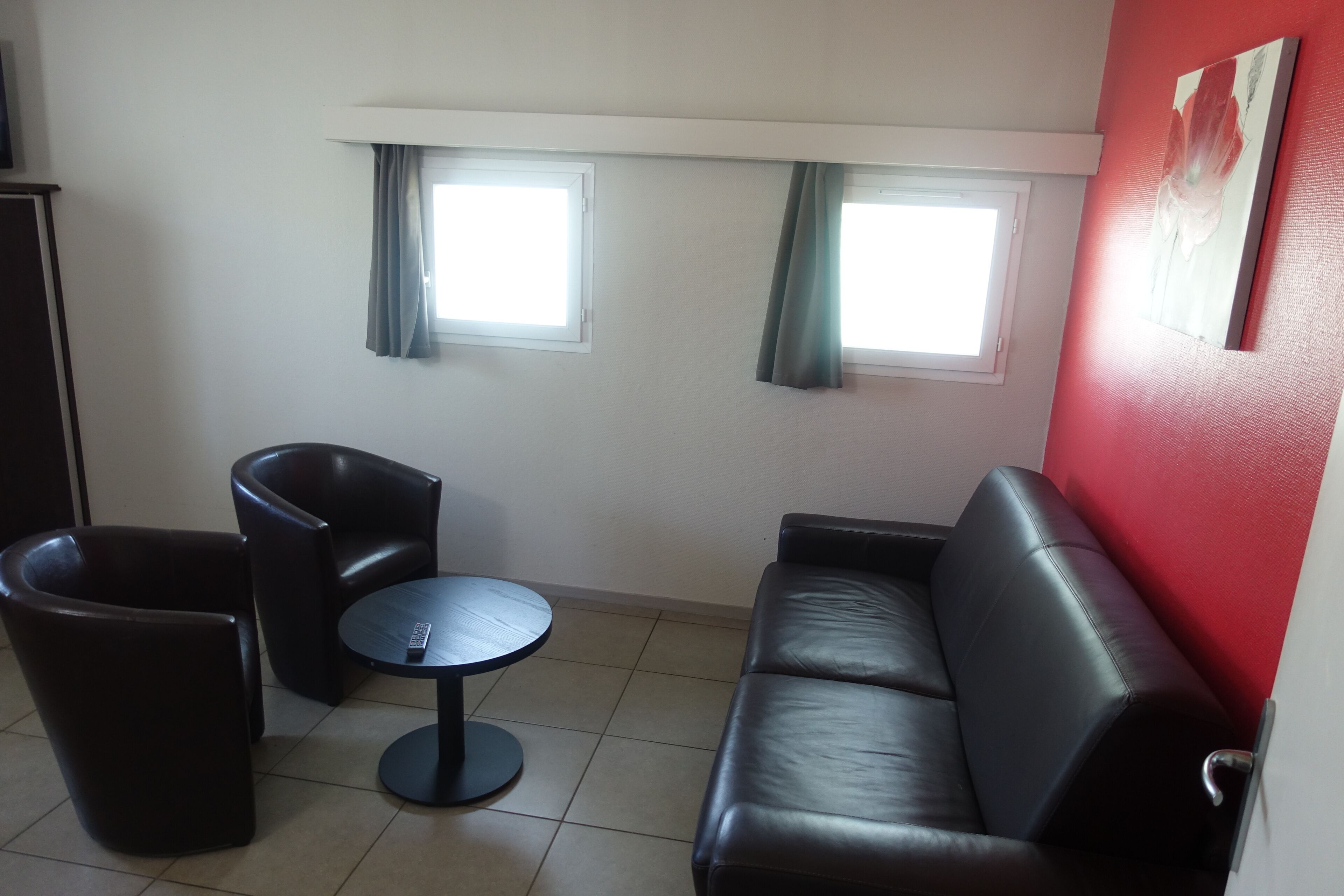 A room with a black leather sofa, a round table, and two black armchairs. There are two windows with curtains, a red accent wall, and a door on the left.
