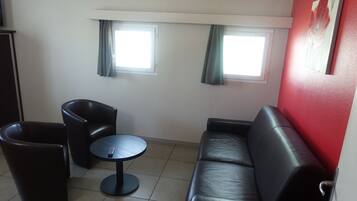 Une pièce avec un canapé en cuir noir, une table ronde et deux fauteuils noirs. Il y a deux fenêtres avec des rideaux, un mur d’accent rouge et une porte sur le côté gauche.
