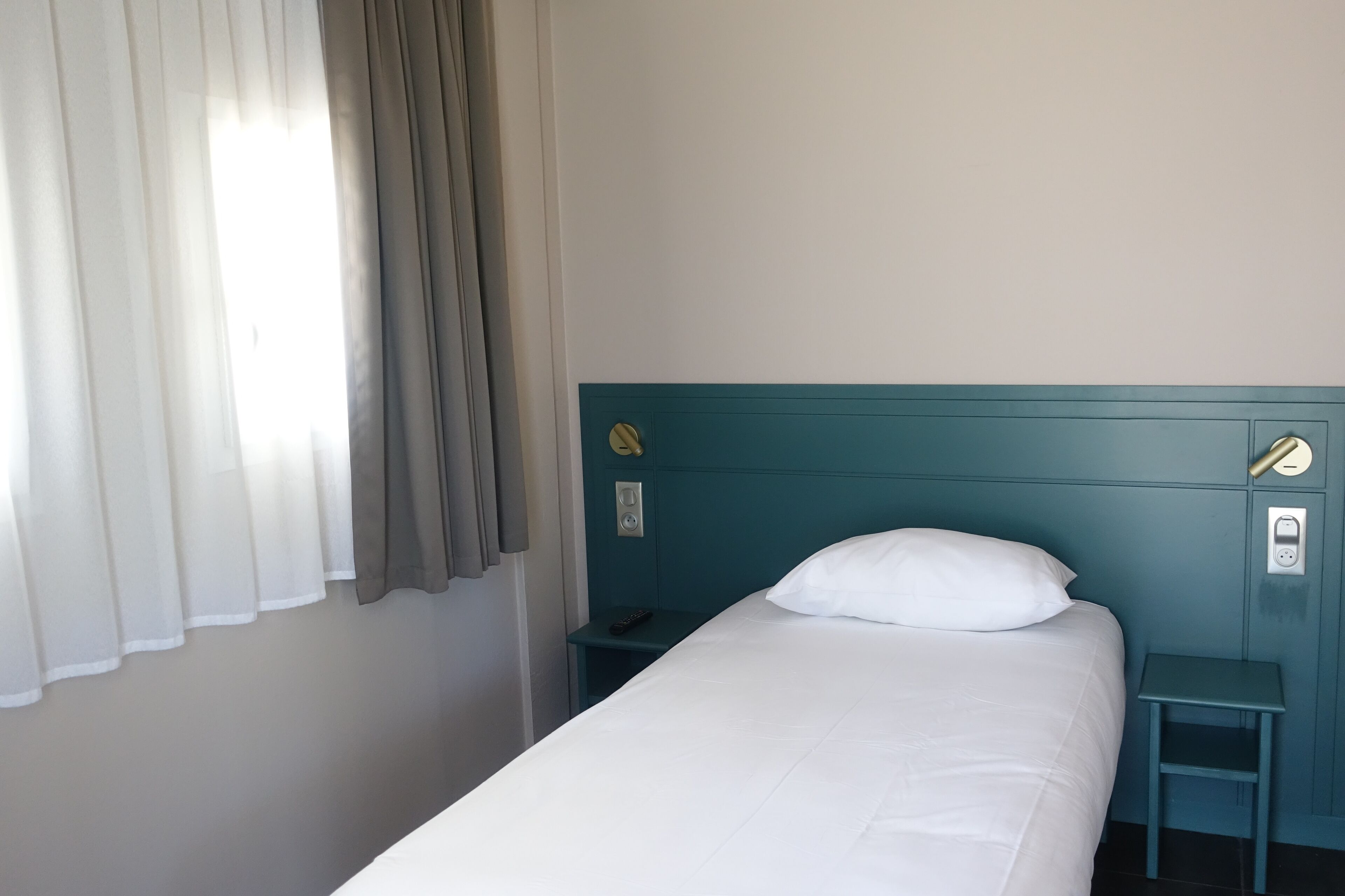 A single bed with a white pillow, a teal headboard, a bedside table with a remote control, and a power outlet on the wall.