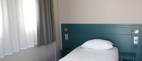 A single bed with a white pillow, a teal headboard, a bedside table with a remote control, and a power outlet on the wall.