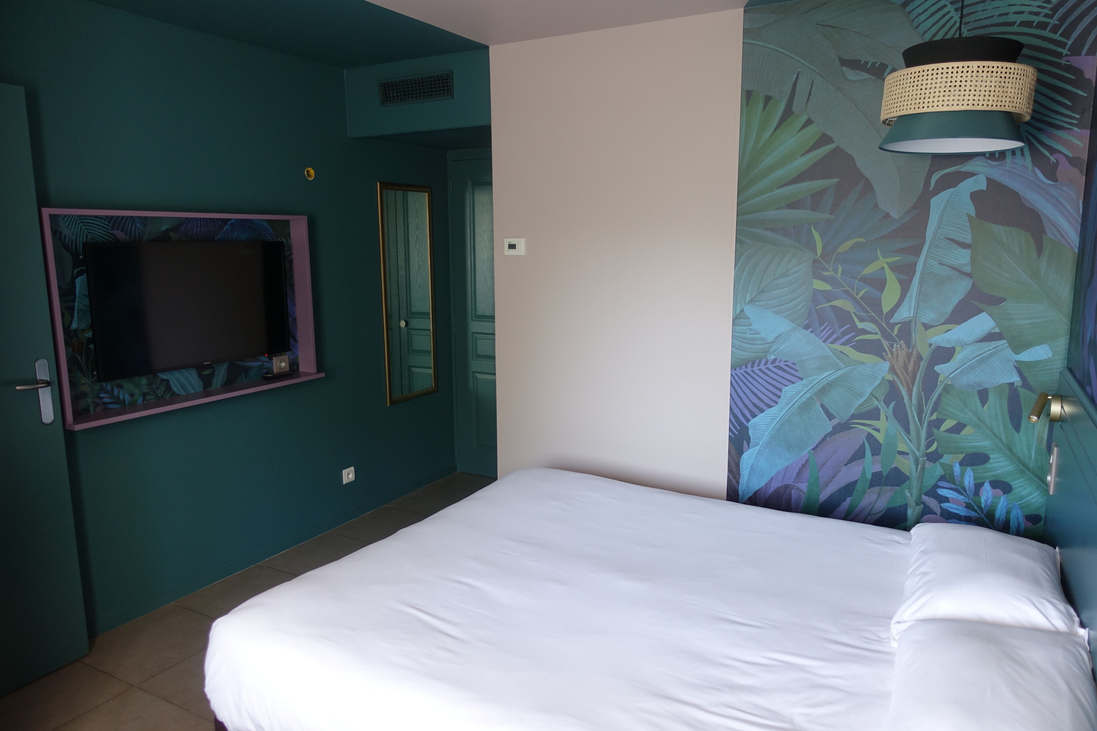 Une chambre d’hôtel avec un lit, une télévision, une porte et un mur orné d’un motif de feuilles tropicales.