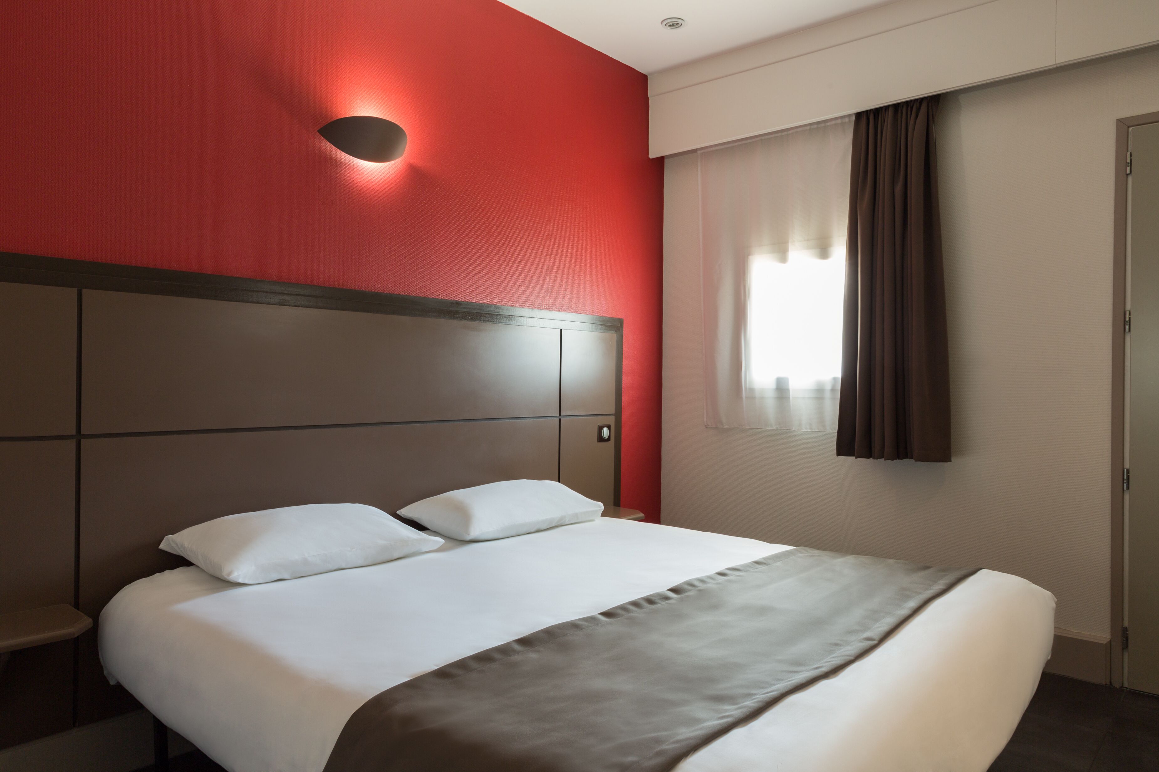 Una habitación de hotel con una cama grande, cabecera, luz fijada en la pared, ventana con cortinas y una puerta.
