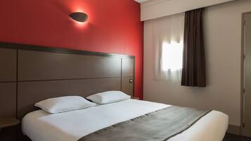 Une chambre d’hôtel avec un grand lit, une tête de lit, une lampe fixée au mur, une fenêtre avec des rideaux et une porte.