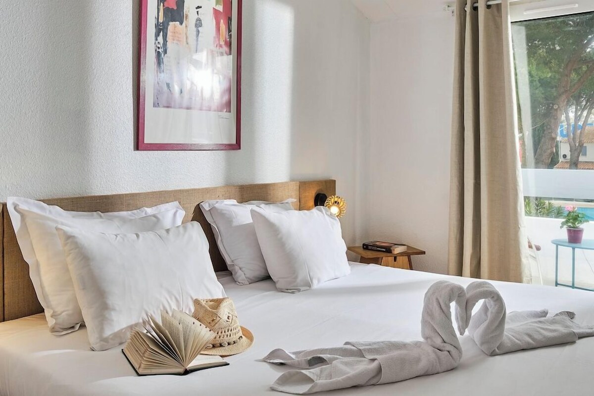 Un cuarto de hotel con una cama grande, ropa de cama blanca, un cuadro en la pared y vistas al exterior por la ventana.