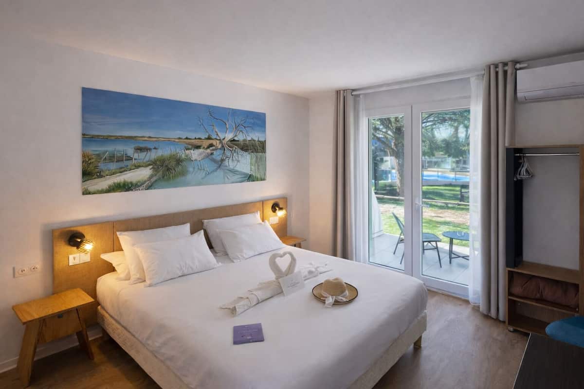 A hotel room with a large bed, a painting on the wall, and a view of a pool and garden through the glass door.