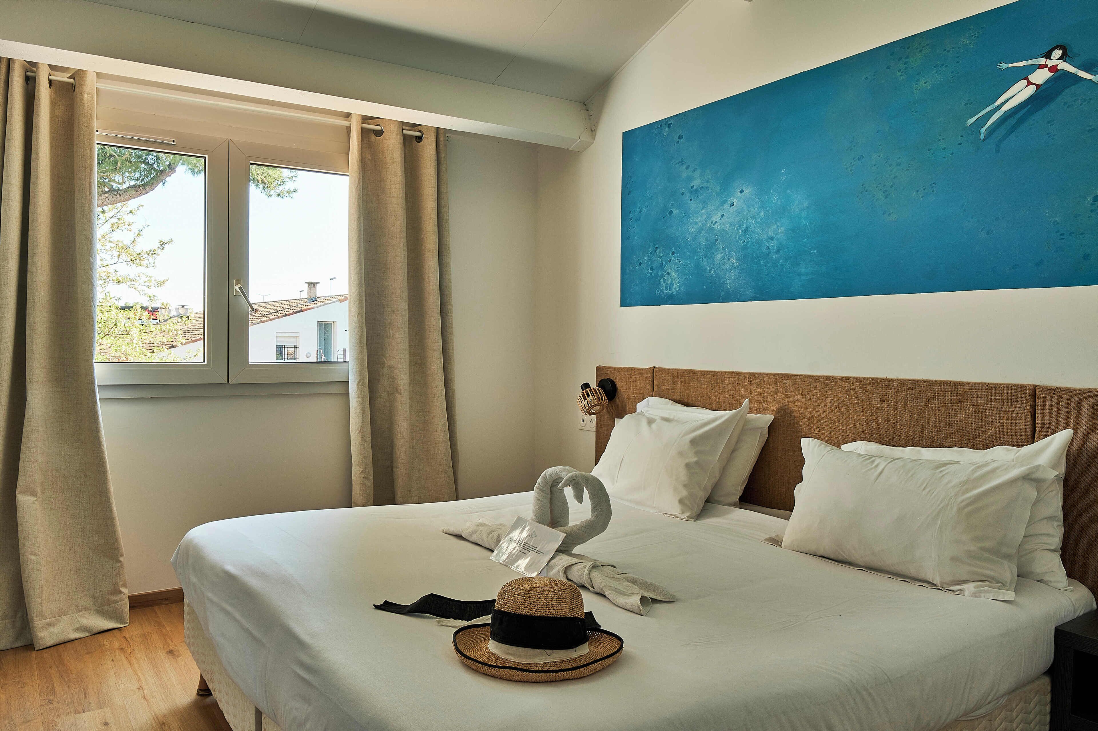 A hotel room with a bed, a window with curtains, a wall-mounted phone, and a painting of a woman swimming.