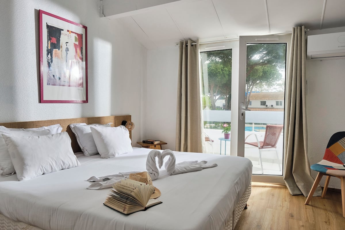 A hotel room with a bed, a framed picture on the wall, a window with curtains, and a view of the outside.