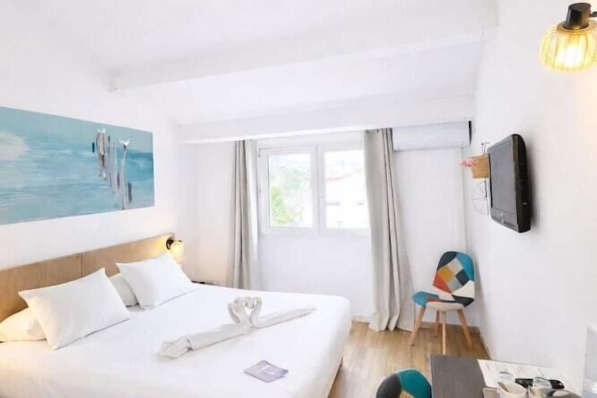 A hotel room with a bed, a desk, a chair, a TV, and a painting on the wall.