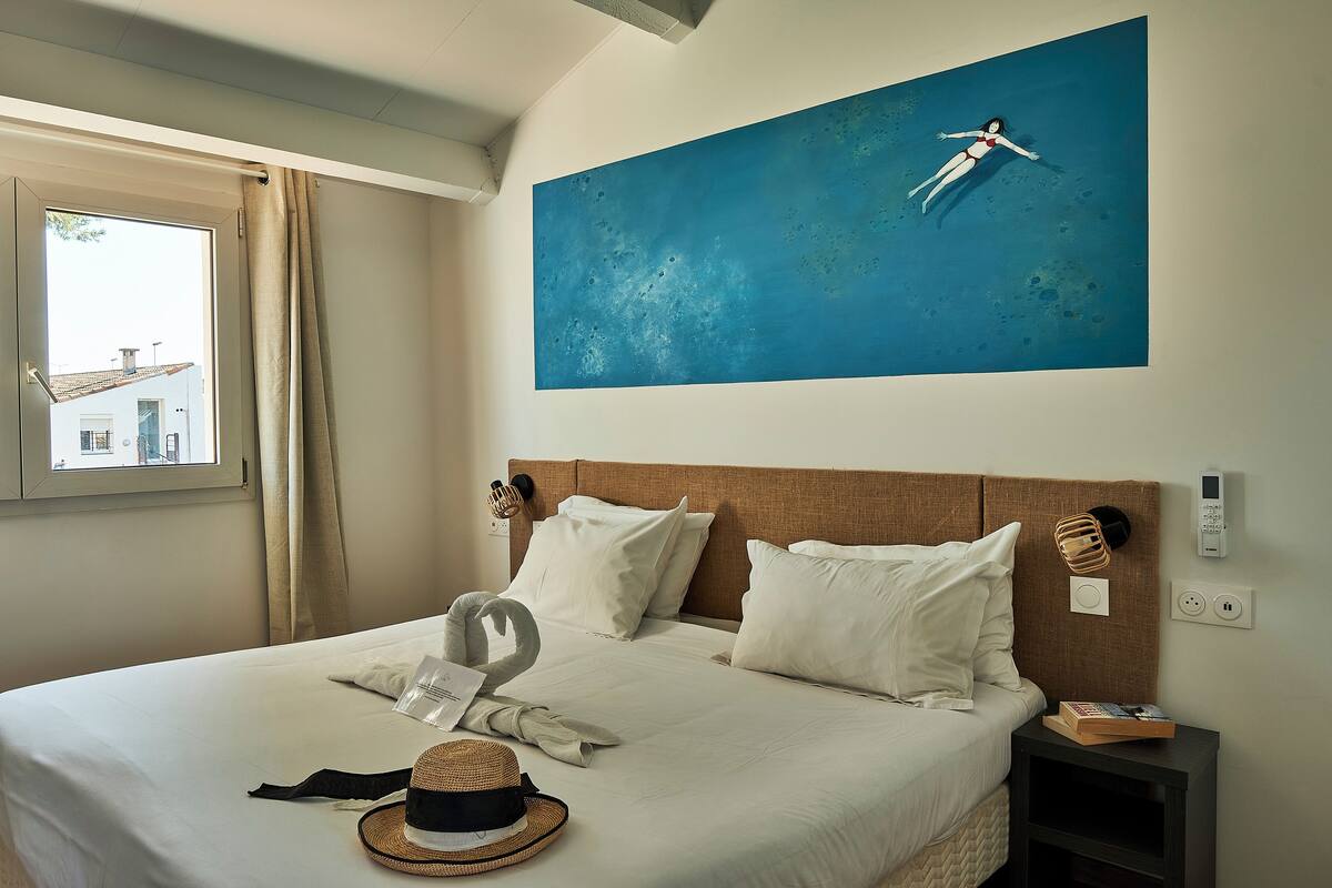 A neatly made bed with white linens, a wall-mounted artwork of a swimmer, and a window with curtains.