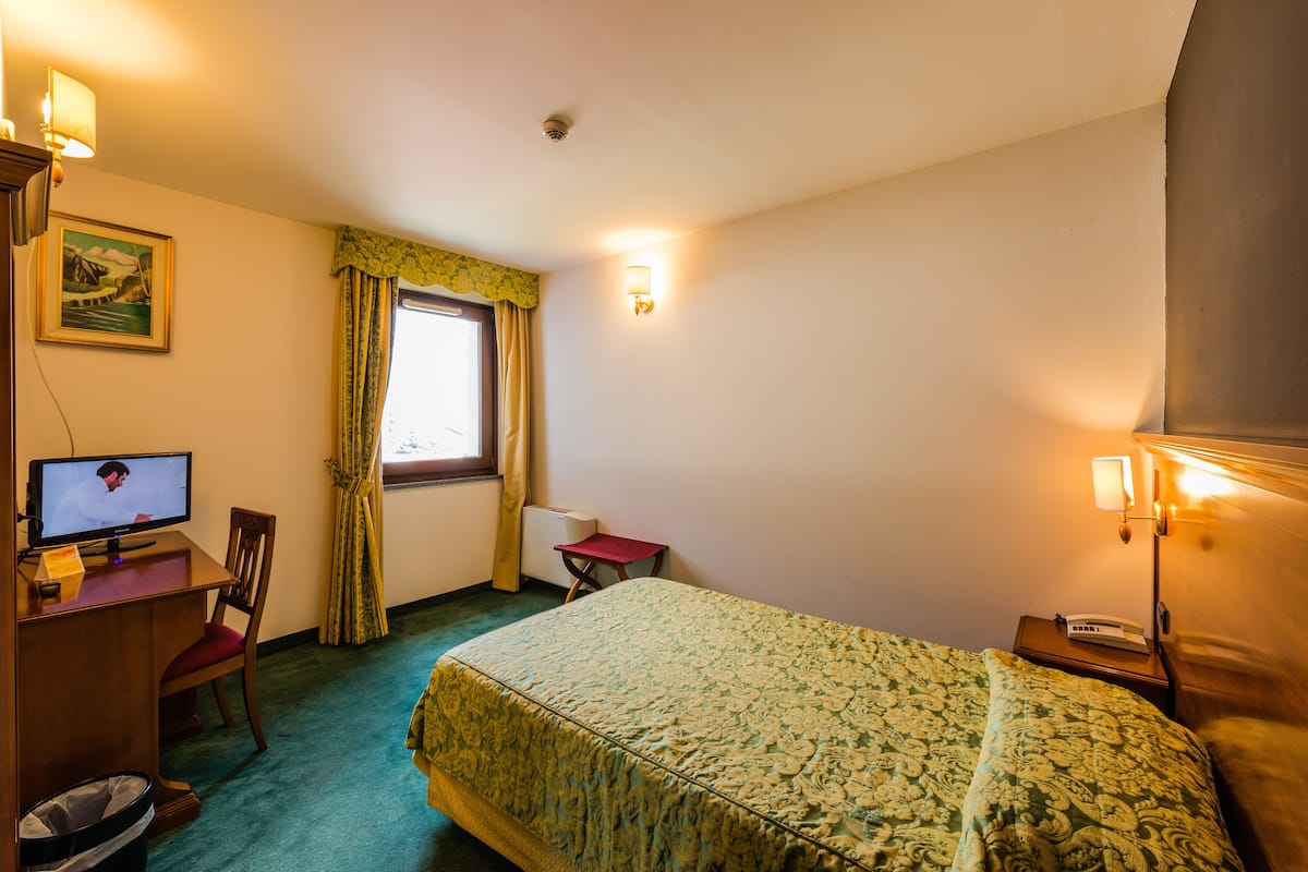 Une chambre d’hôtel comprenant un lit, un bureau avec un ordinateur, une chaise, une fenêtre avec des rideaux et une lampe fixée au mur.