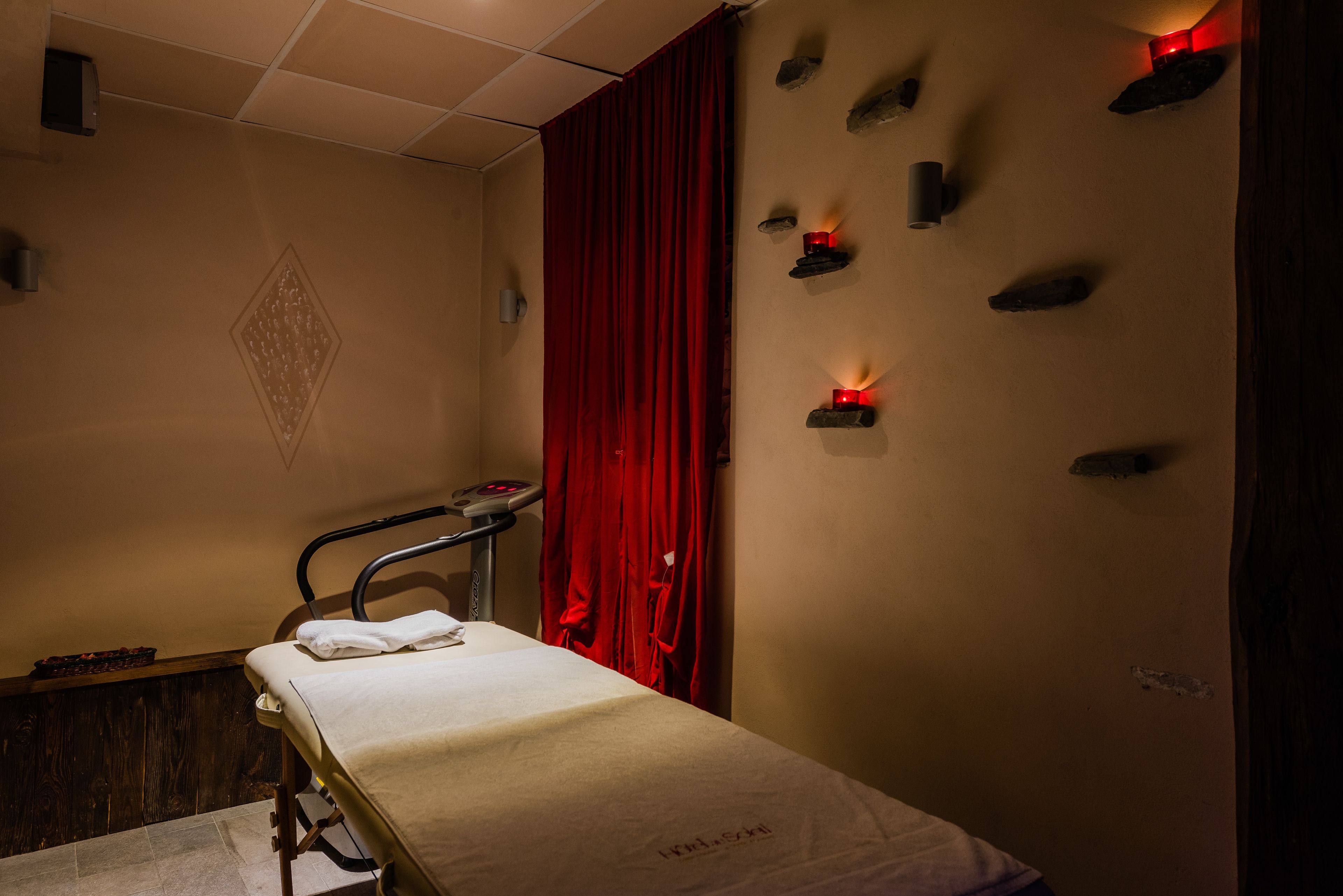 Un cuarto de tratamientos con una cama, una camilla de masaje, una cortina roja y velas fijadas a la pared.