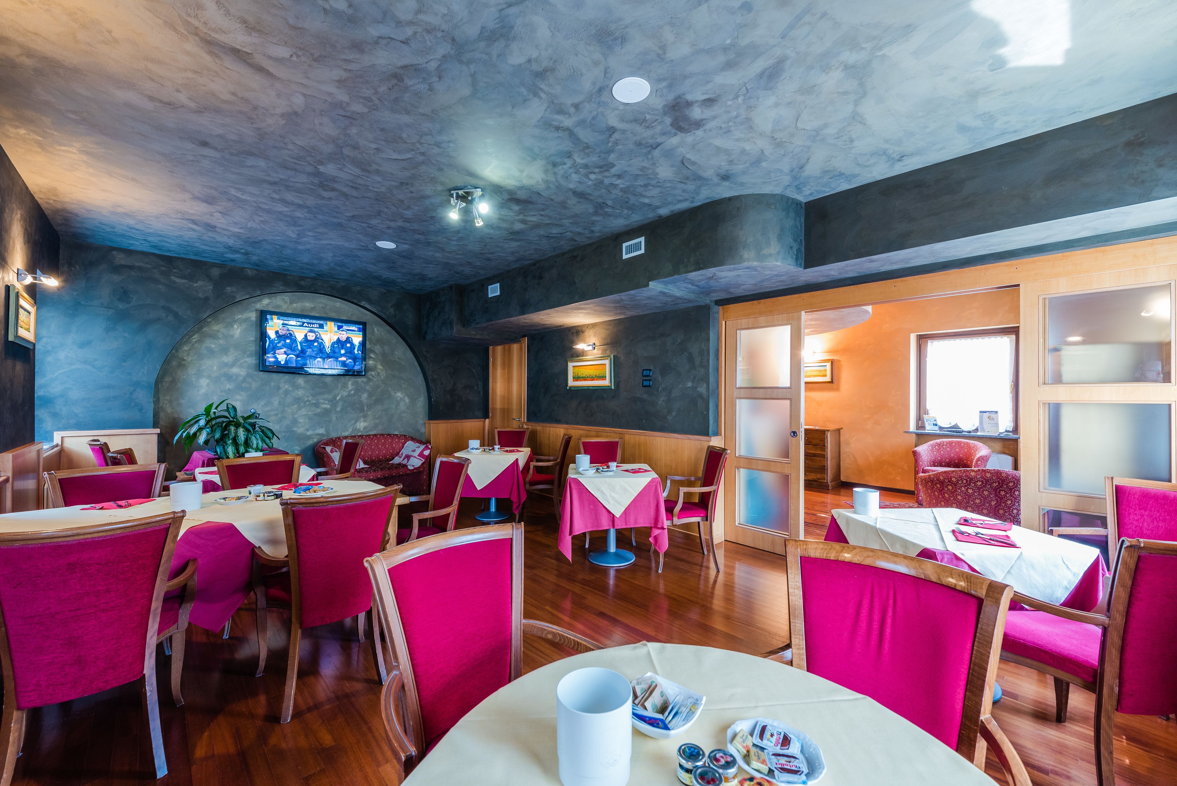 Ein Restaurant mit runden Tischen und roten Stühlen, einem an der Wand montierten Fernseher und einer Holzbalkendecke.