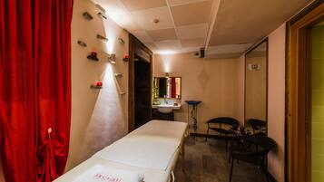 A room with a bed, a small table, and a chair. There is a window with curtains, a sink, and a mirror.