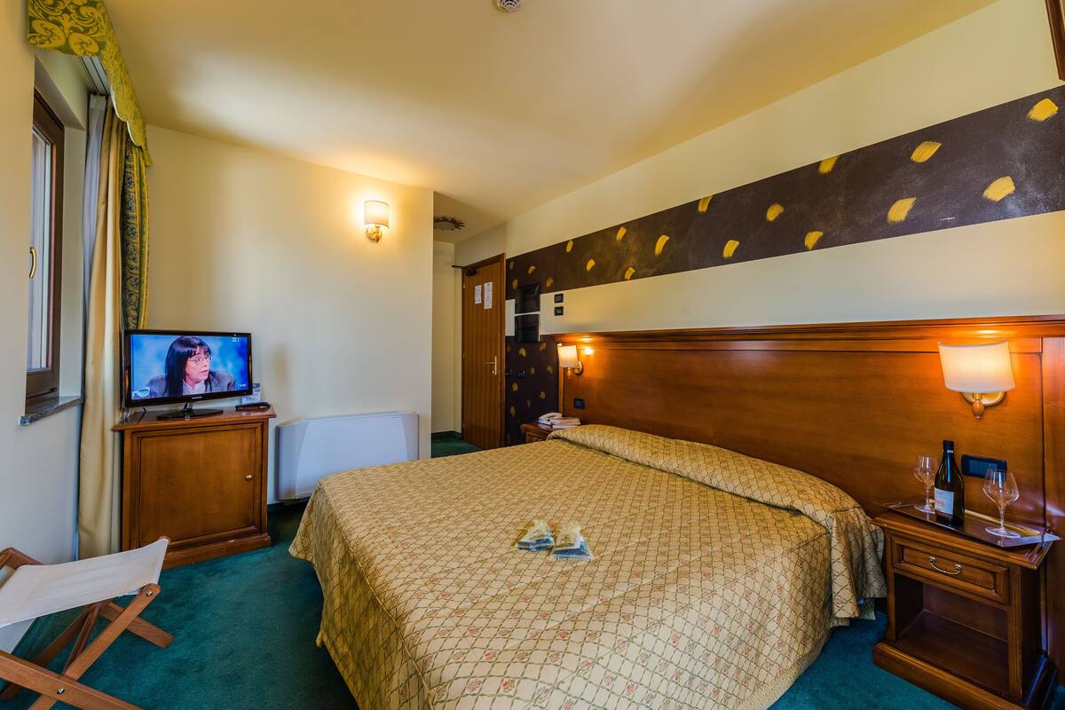 Une chambre d’hôtel avec un lit, une télévision, une tête de lit en bois et une table de chevet sur laquelle se trouvent une bouteille et des verres.
