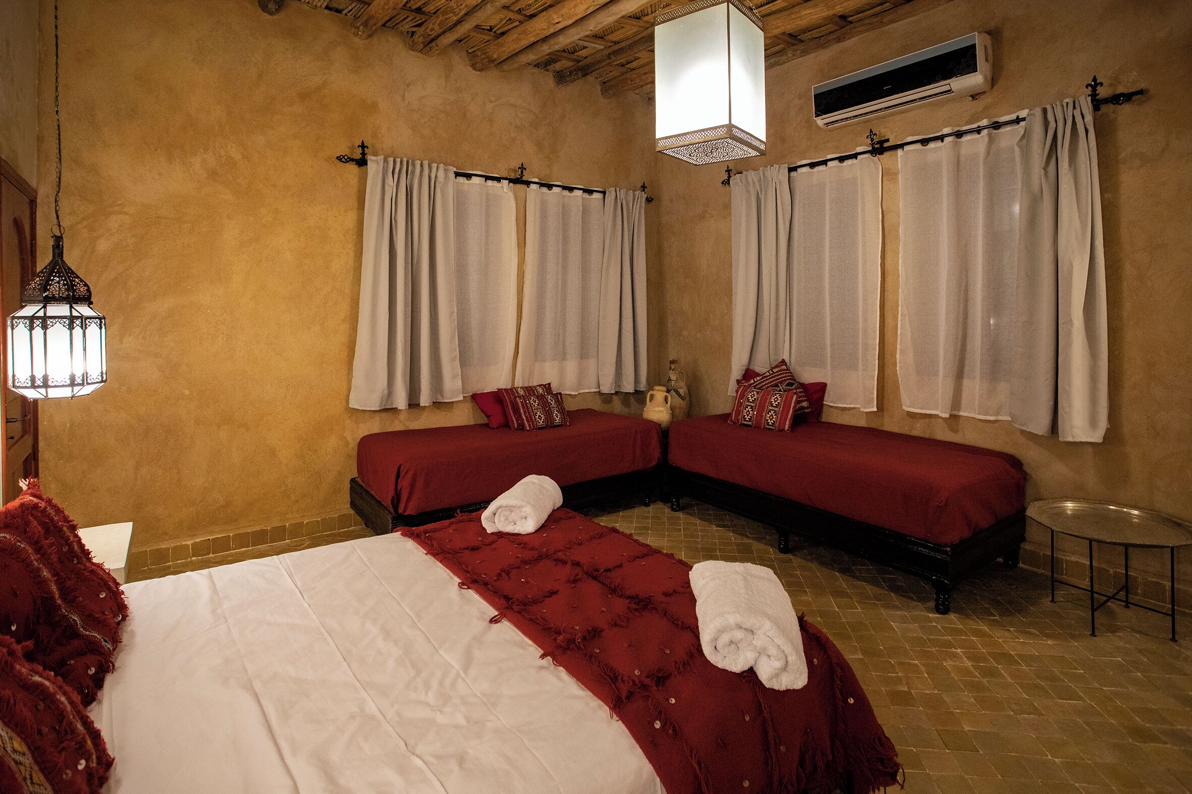 A hotel room with two single beds, a large bed, and a small table. There are two windows with curtains, a hanging light fixture, and an air conditioning unit on the wall.