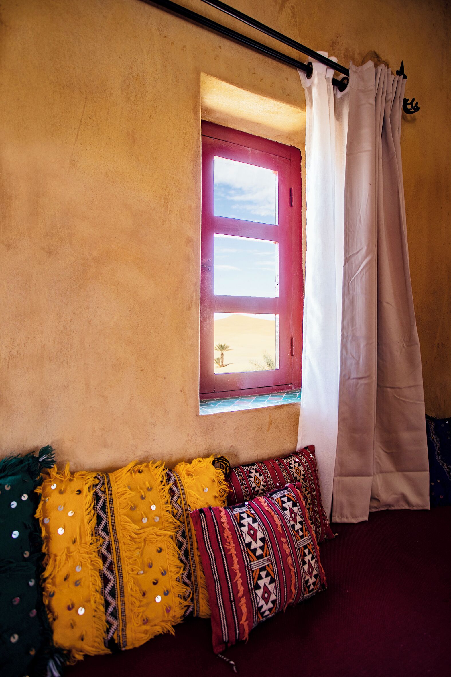 A room with a window, a red door, and a curtain. There are colorful pillows on a bench.