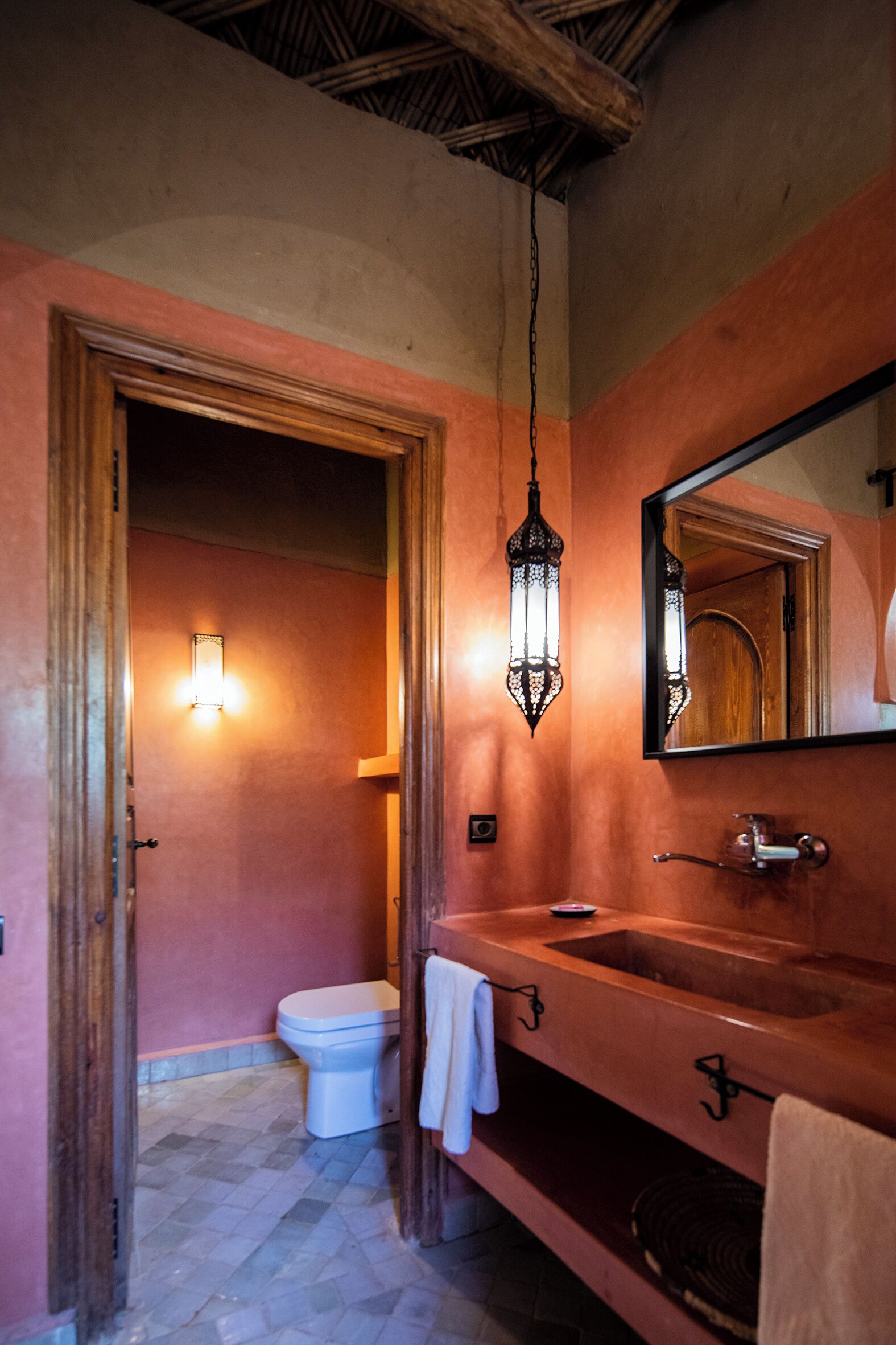 A bathroom with a wooden door, a white toilet, a large mirror, and a hanging light fixture.