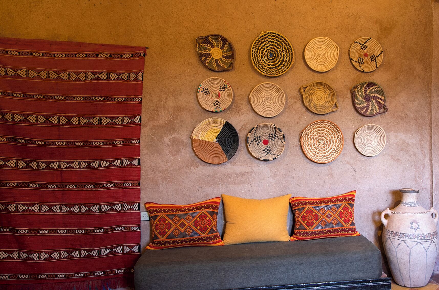 Una habitación con una cama, almohadones estampados y un muro decorado con tapetes tejidos.