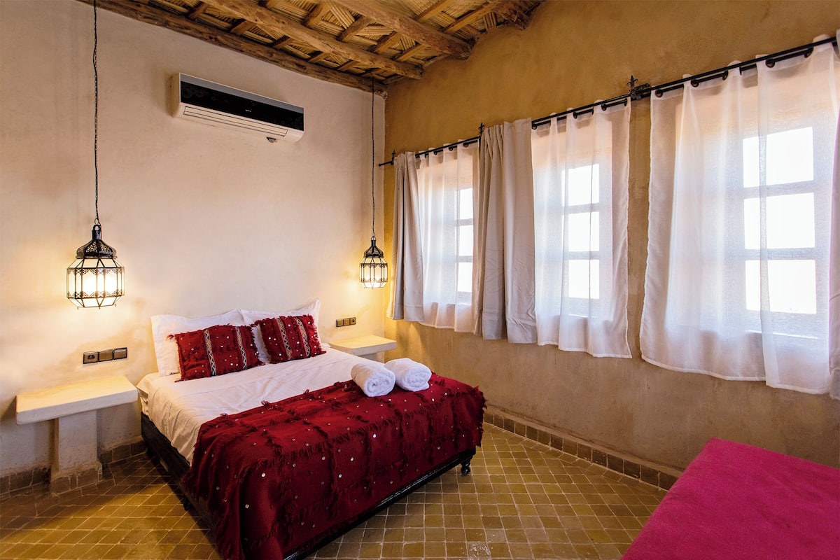 Un dormitorio con una cama, dos mesitas de noche, una lámpara colgante, un ventanal con cortinas y un suelo de baldosas.