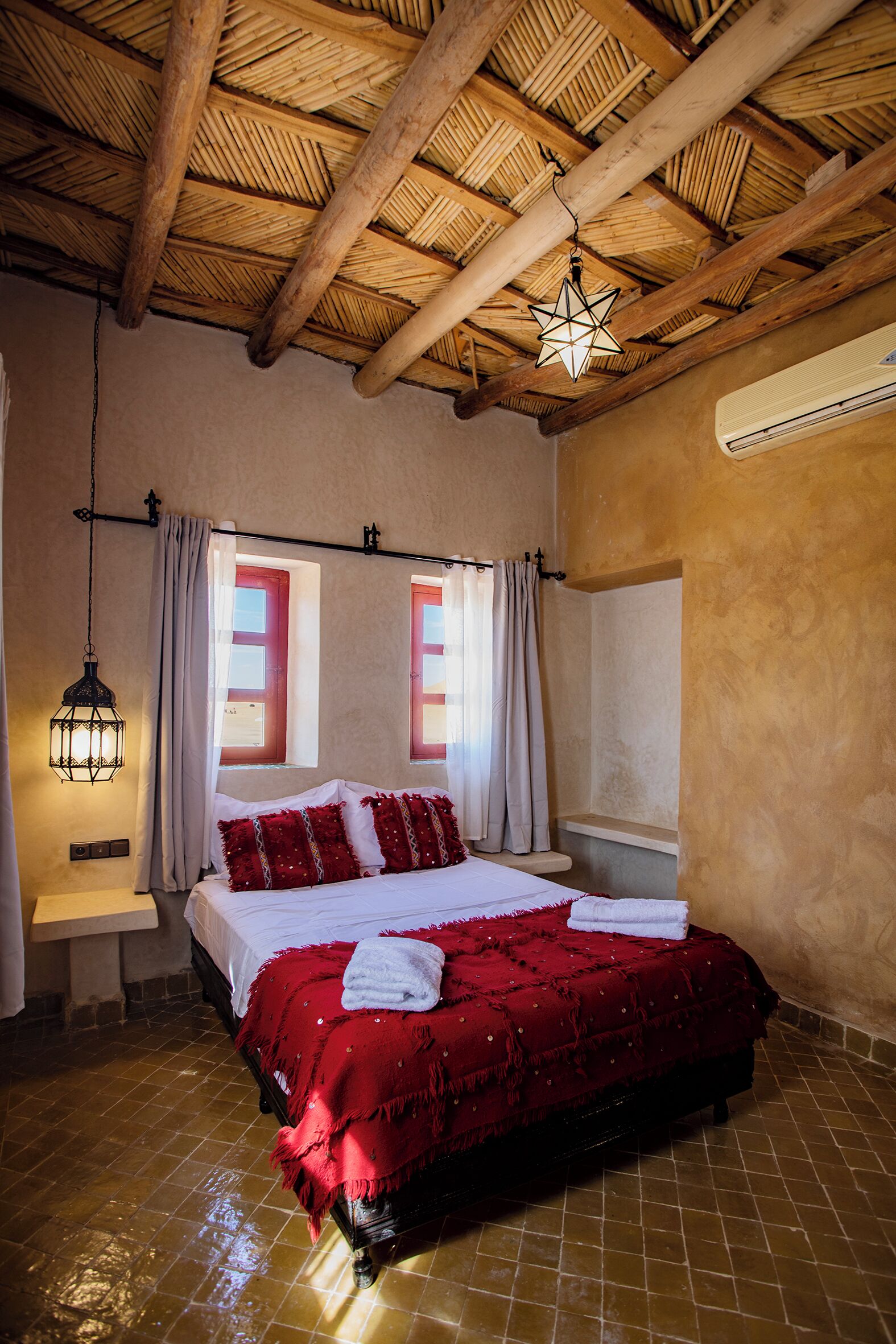 A bedroom with a wooden ceiling, a bed with a red bedspread, two windows with curtains, and an air conditioning unit on the wall.