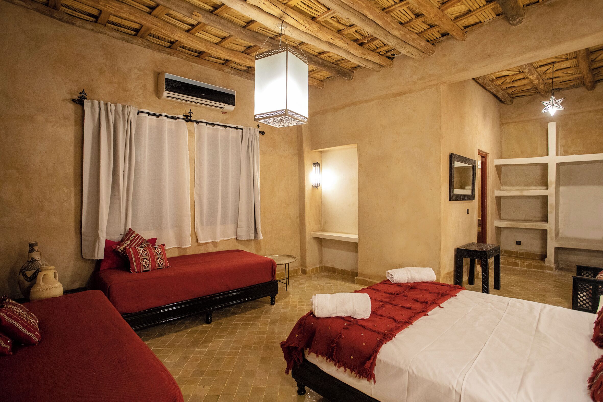 A room with two beds, a wooden ceiling, a wall-mounted air conditioner, and a small table with a vase.