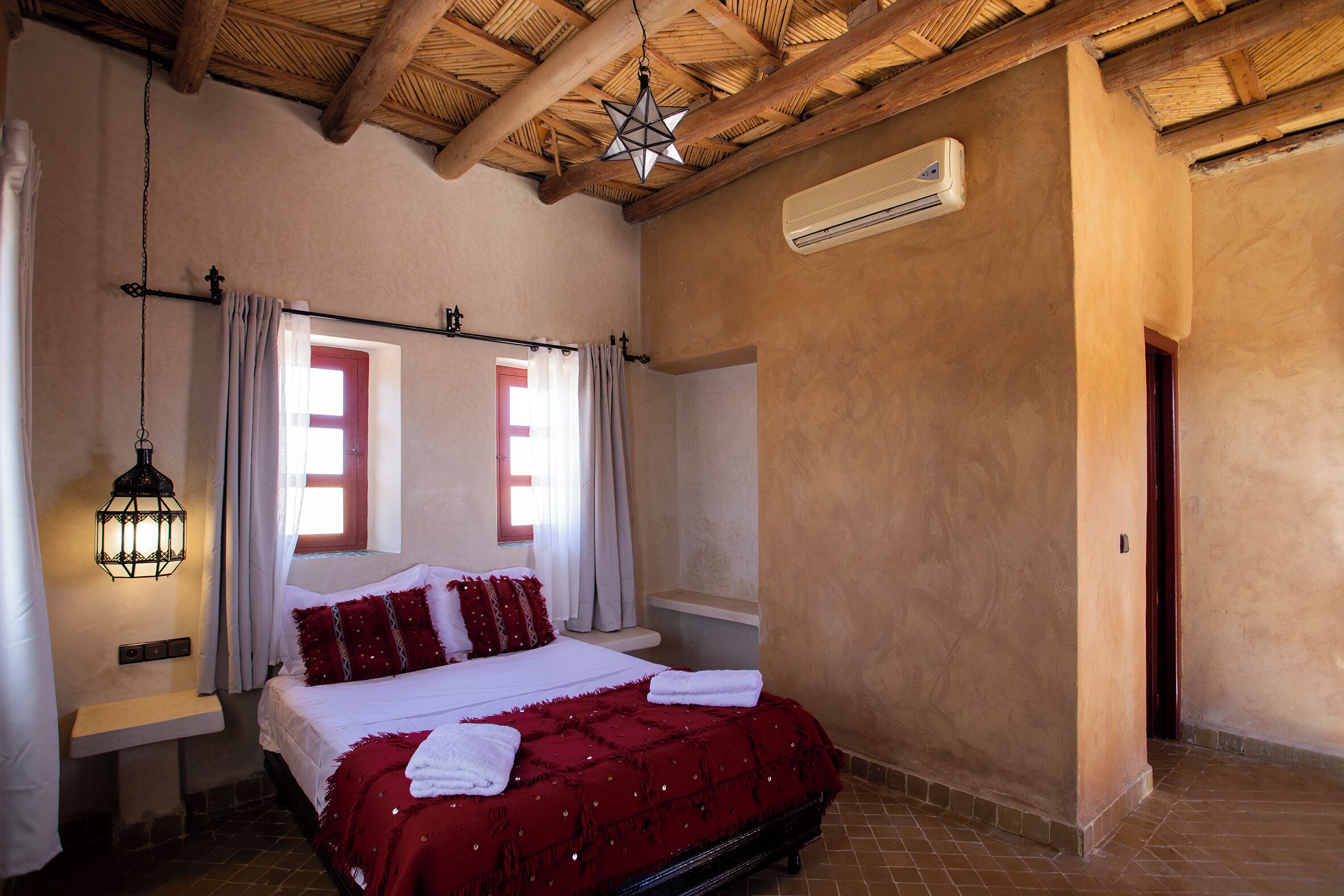 Un dormitorio con techo de madera, una cama con ropa de cama roja, una mesita de noche, un farol colgante y una ventana con cortinas.