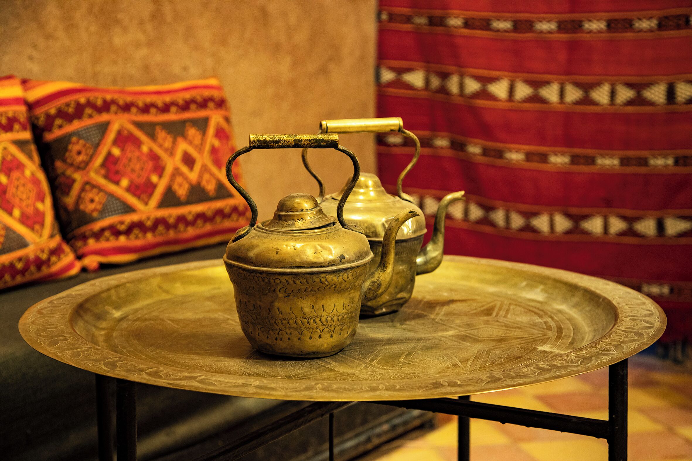 Un plateau en laiton sur lequel reposent deux théières décorées, placé sur une table. L’ensemble est mis en valeur par un coussin à motifs et un fond en tissu rouge.