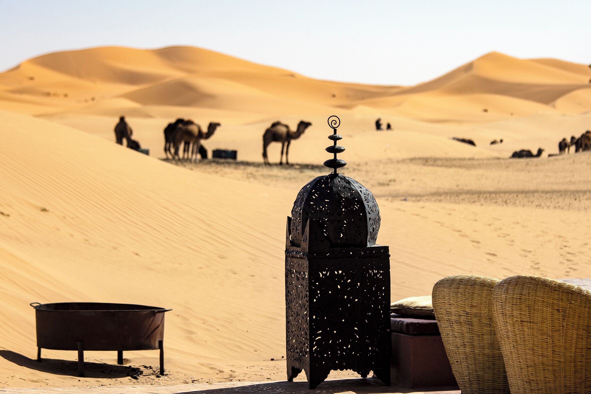 A desert scene with sand dunes, a traditional lantern, a metal pot, and a woven chair.
