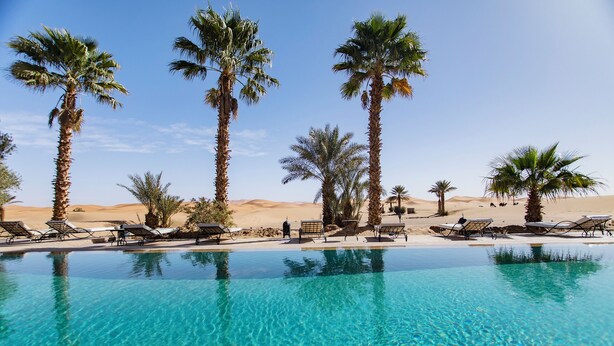 A clear pool surrounded by palm trees and lounge chairs, with a desert landscape in the background. - Hotel Kanz Erremal (Taouz)