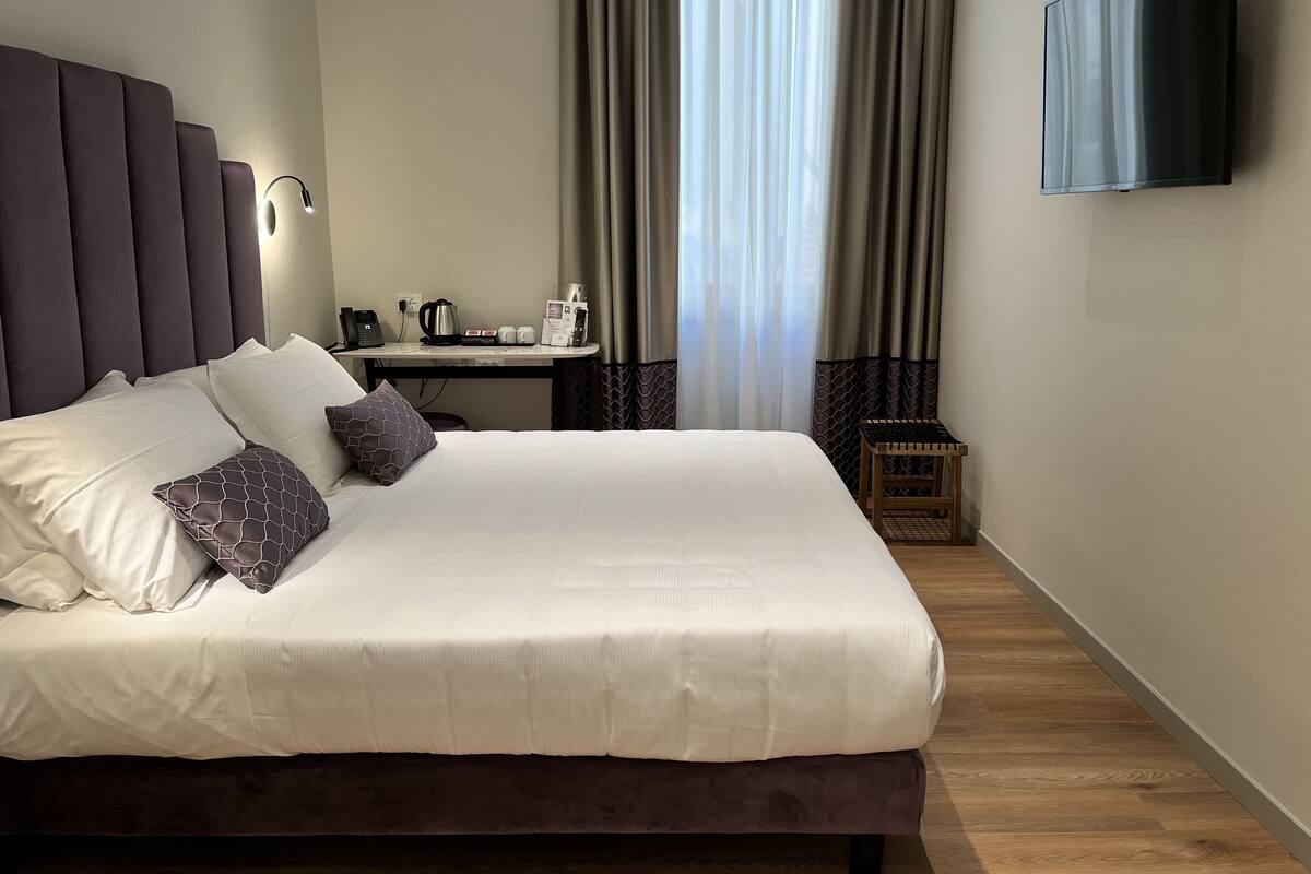 Een netjes opgemaakt bed met witte en gemêleerde kussens, een nachtkastje met een koffiezetapparaat en kopjes, en een televisie aan de muur.
