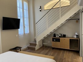 Camera d'albergo moderna con pavimento in legno, un letto, una televisione a parete, una scala con ringhiera, un tavolino e un armadietto in legno con elettrodomestici.