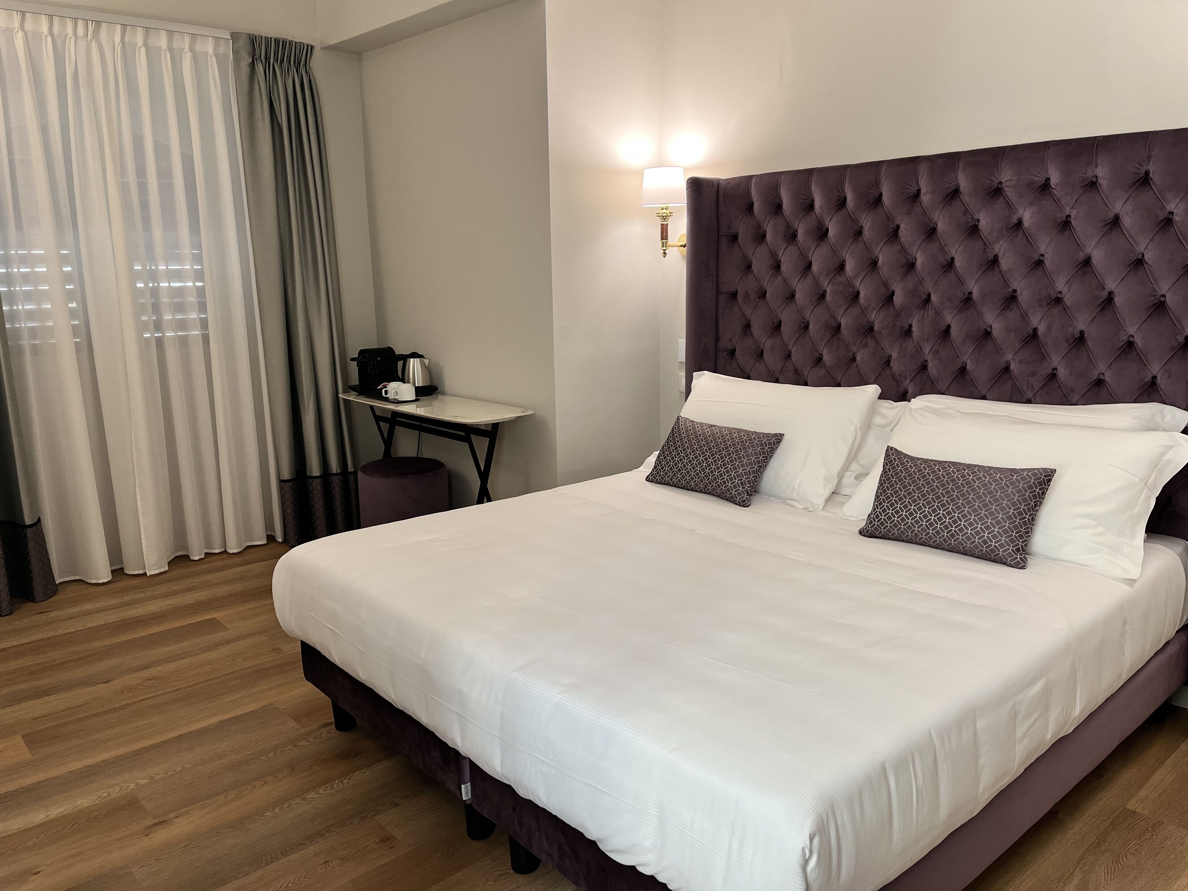 A hotel room with a large bed, a bedside table with a coffee maker, and a wall-mounted light.