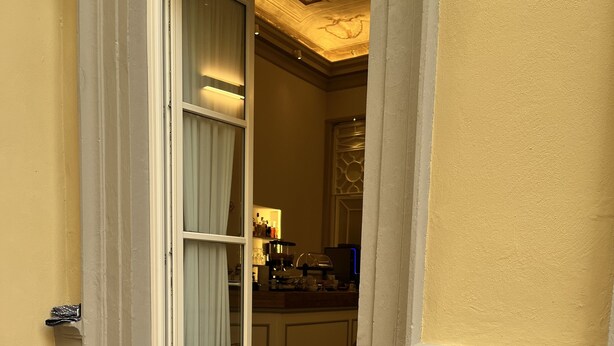 Habitació d'hotel amb taula de menjador preparada per a dues persones, una cadira i vista a la cuina i la zona del bar a través d'una porta oberta.