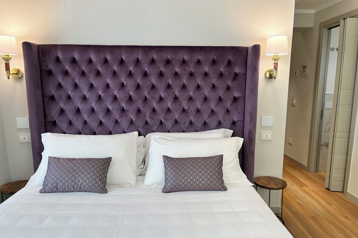 Una cama con cabecera capitonada, ropa de cama blanca y almohadas estampadas.