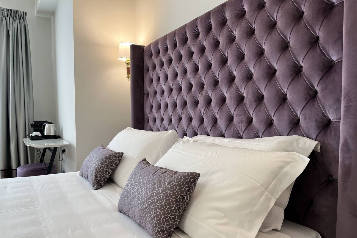 Een hotelkamer met een gestoffeerd hoofdeinde, een bed met witte en gemêleerde kussens, een nachtkastje met een lamp en een koffiezetapparaat.