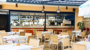 Een terrasrestaurant met tafels en stoelen, een bargedeelte met verschillende flessen, en een stenen vloer.