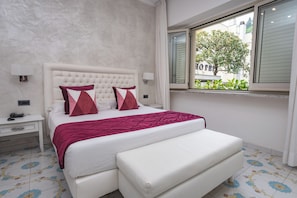 A bedroom with a large bed, a bedside table, a nightstand, a window with a view of a building, and a patterned floor.