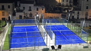 Een tennisbaan buiten met een metalen hek, omringd door gebouwen.