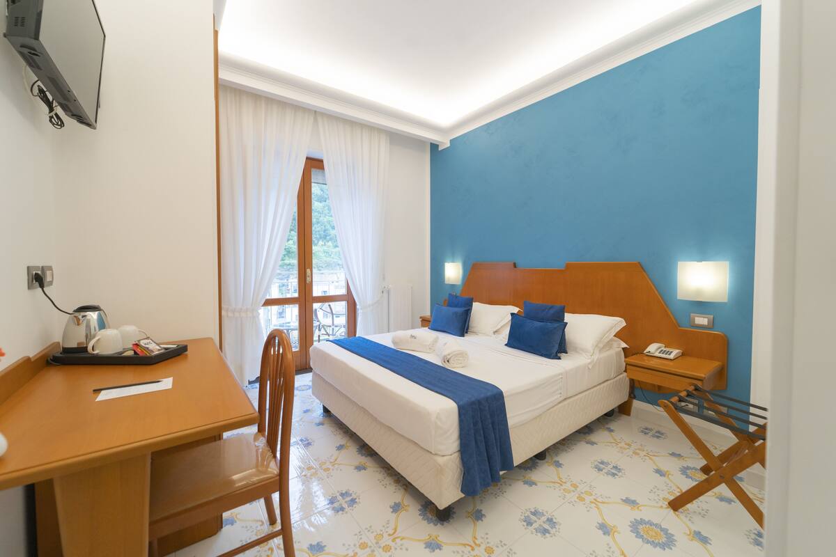 Une chambre d’hôtel avec un lit, un bureau, une chaise, une table de chevet et une fenêtre avec des rideaux.