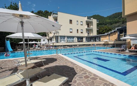 A swimming pool with lounge chairs and umbrellas, a building with balconies, and a mountainous backdrop.
